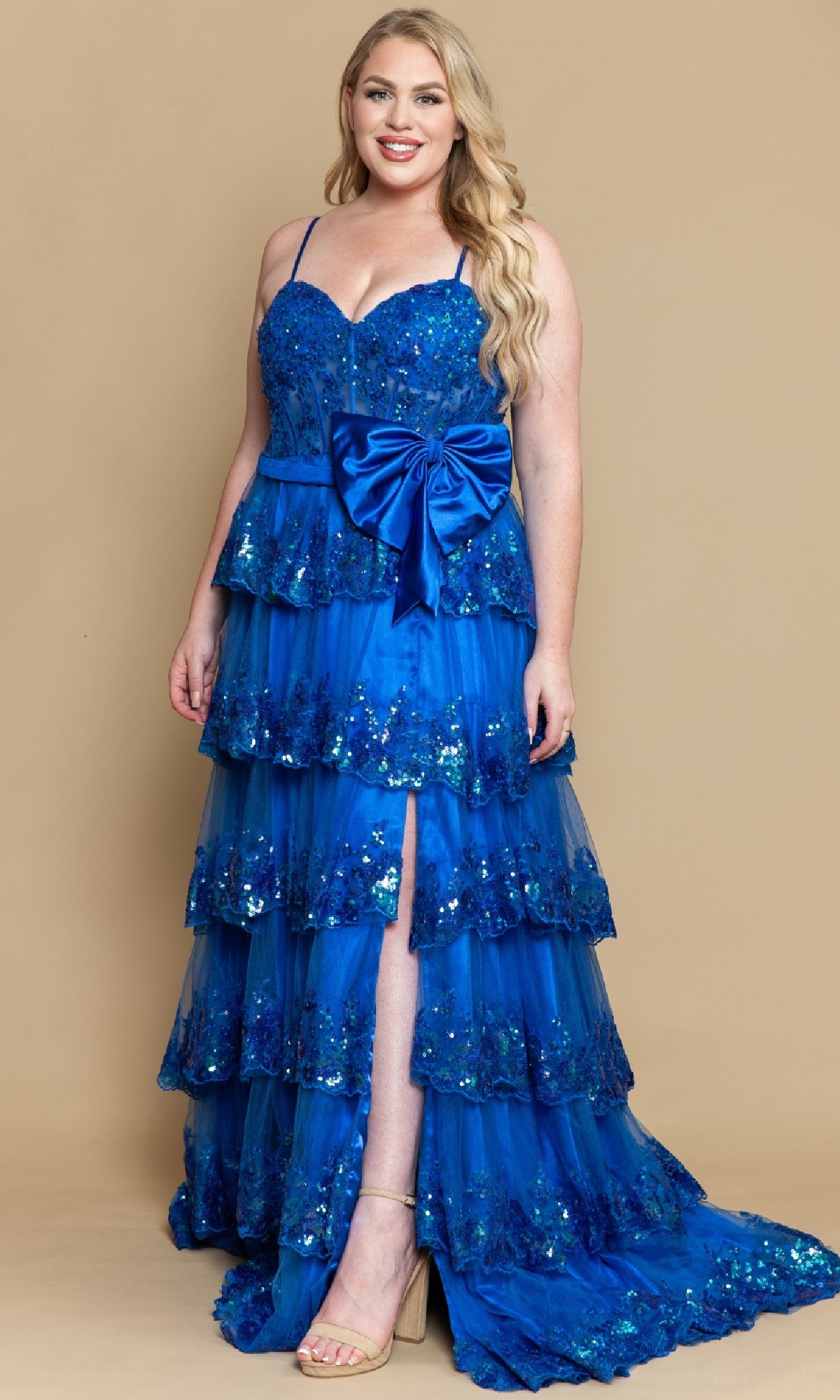 A woman wears the PolyUSA W1162 Plus-Size Tiered Prom Dress with sequins, spaghetti straps, a large waist bow, and a front slit, standing against a plain tan background.